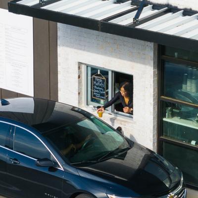 Server at drive-thru window handing beverage to customer in a dark-colored car.