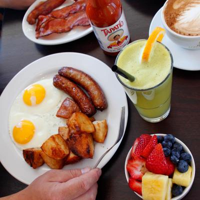 Fried eggs with sausage and potatoes accompanied by mixed fruit, bacon, and a fresh juice.