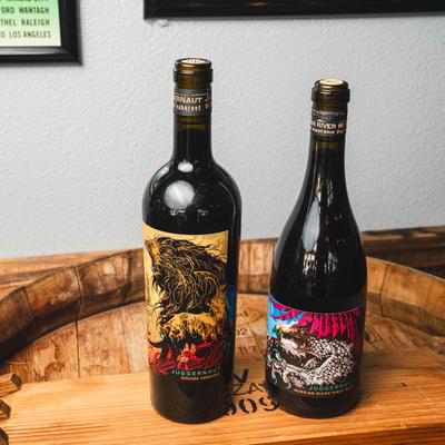 Two bottles of wine on a barrel head by a white wall.