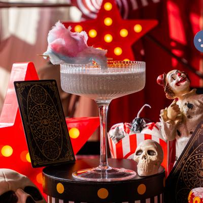 Coupe cocktail topped with cotton candy, set against a Halloween-themed circus.