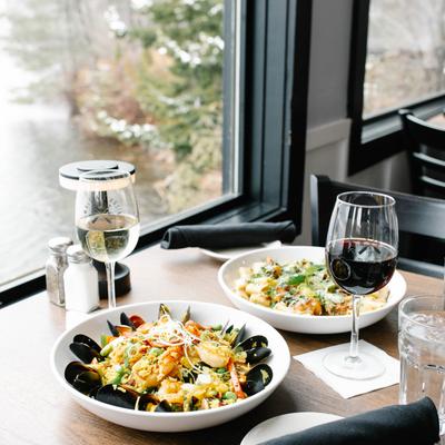 A table by a window overlooking a lake, set with seafood paella, pasta, wine, and glasses of water.