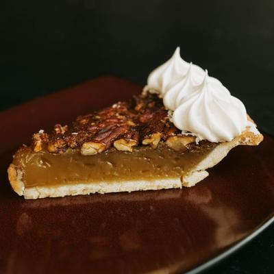 A slice of pecan pie topped with whipped cream on a dark brown plate..