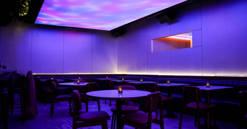 Interior of a lounge with purple and pink ceiling lights, dark tables, chairs and candles on tables