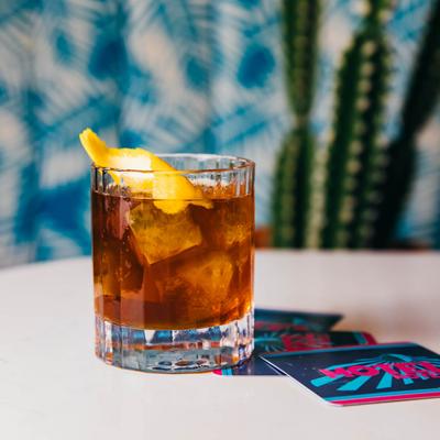 A tumbler with dark liquid and ice, garnished with lemon peel, sits on a table.