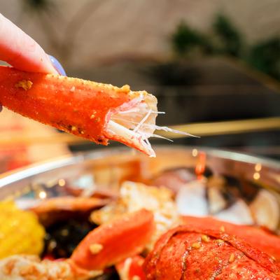 Hand holding a crab leg above the bowl of seafood boil, close-up.