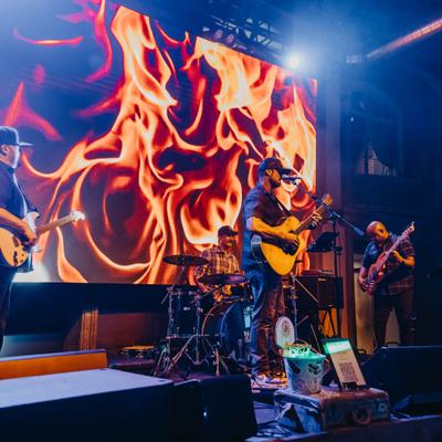 Live band performing on stage with a large video screen displaying abstract flames.