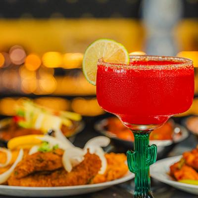 A frosty red margarita sits in a glass with a cactus-shaped stem, beside plates of food.