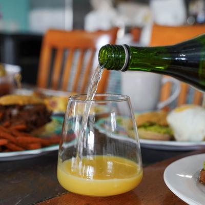 A green bottle pouring sparkling wine into a glass partially filled with orange juice.