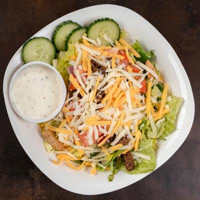 Garden salad with greens, shredded cheese, cucumber slices, and a cup of dressing.