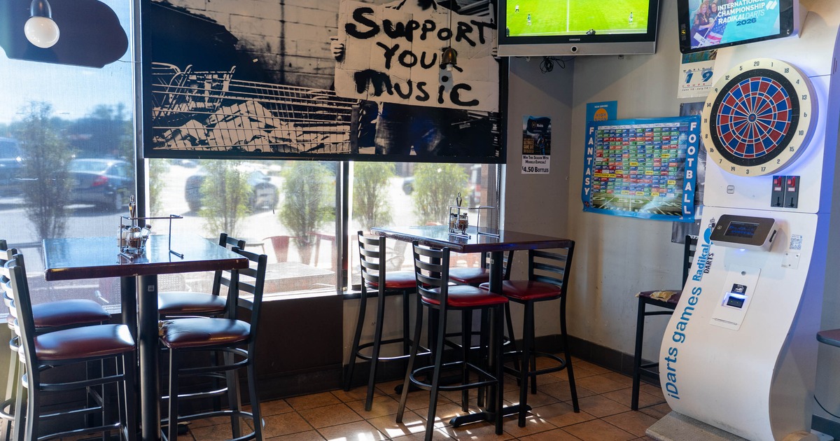 Interior of a bar with tables, chairs, a mural, a dartboard machine, and a TV showing a soccer match