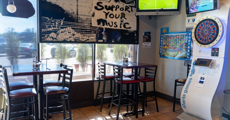 Interior of a bar with tables, chairs, a mural, a dartboard machine, and a TV showing a soccer match