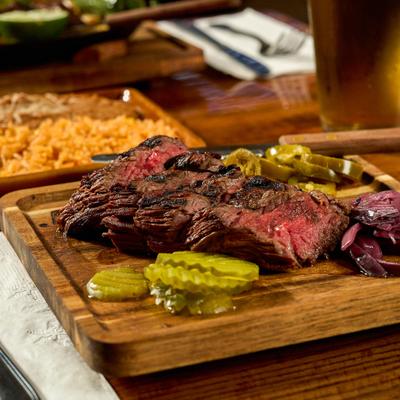 Carne Asada served on a wooden board with pickled vegetables and sides of rice and beans.