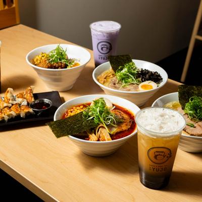 A wooden table with bowls of ramen, a chicken bowl, pork gyoza, and boba teas.