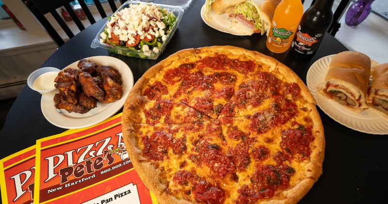 Table spread featuring a whole pizza, wings, Greek salad, sandwich and bottles of soda