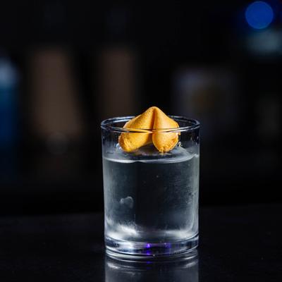 A transparent drink garnished with a fortune cookie, sits on a dark surface.