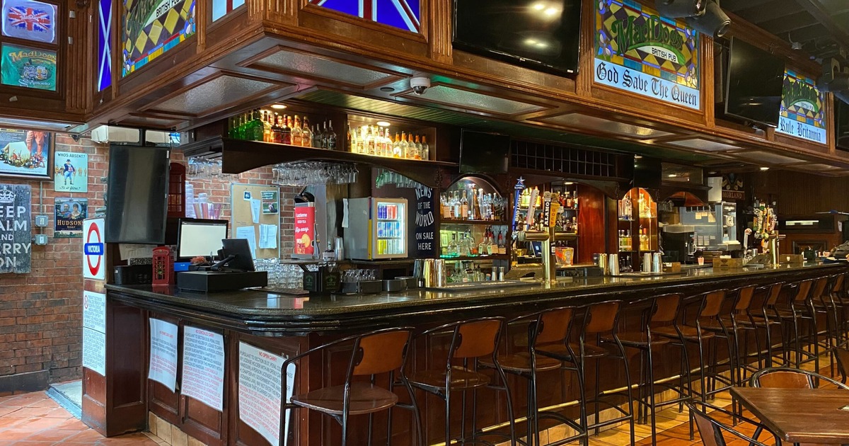 Pub interior with a long wooden bar, high stools, and colorful stained glass decor