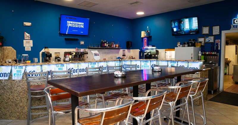 Interior, high table with bar stools, bar in the back, TVs on the walls, kitchen entrance