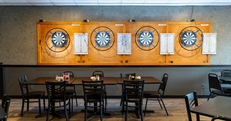 Four dartboards on a wall above two tables and chairs in a recreational room.