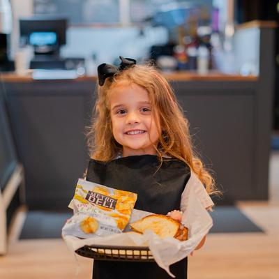 A child holding a basket with a grilled sandwich and a bag of chips.