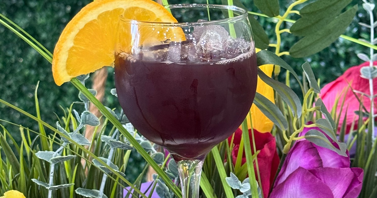 A dark purple cocktail with ice and an orange slice among flowers