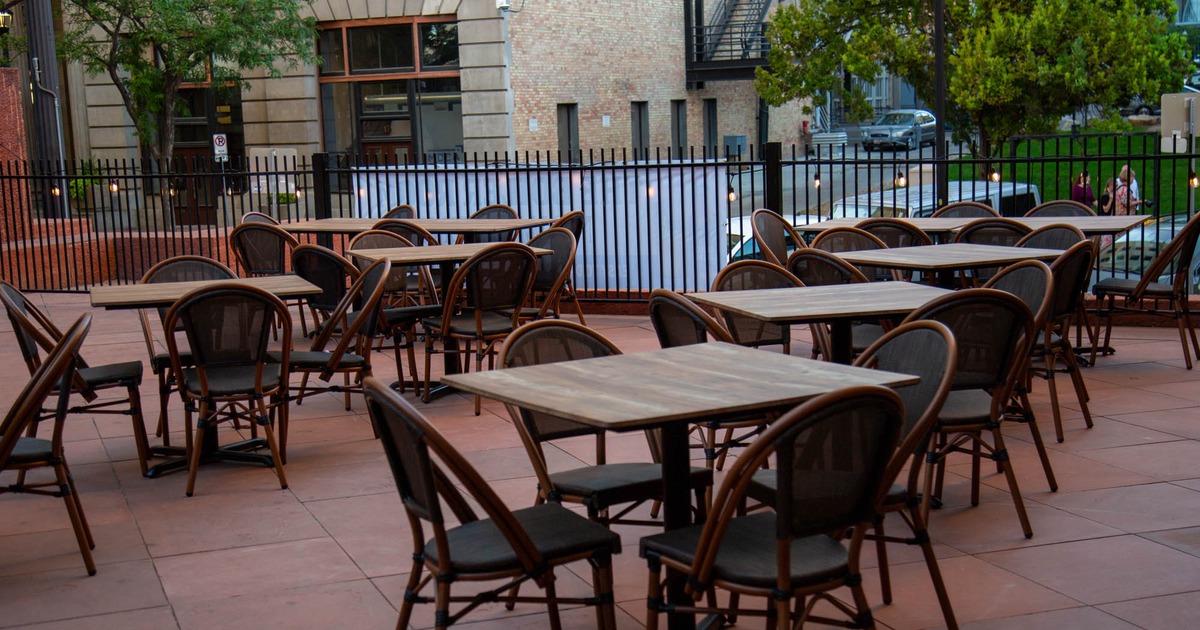Outdoor patio with wooden tables and brown chairs, framed by a black fence, with an urban backdrop