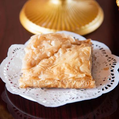 A single piece of baklava resting on a white plate surrounded by golden serve-ware.