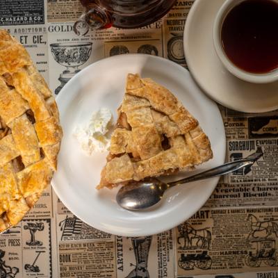 Piece of apple pie with whipped cream, served alongside whole pie and a cup of tea.