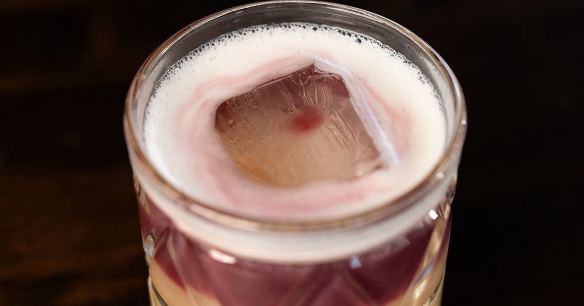 Close-up of a cocktail glass featuring a large ice cube, white frothy top, and a hint of purple