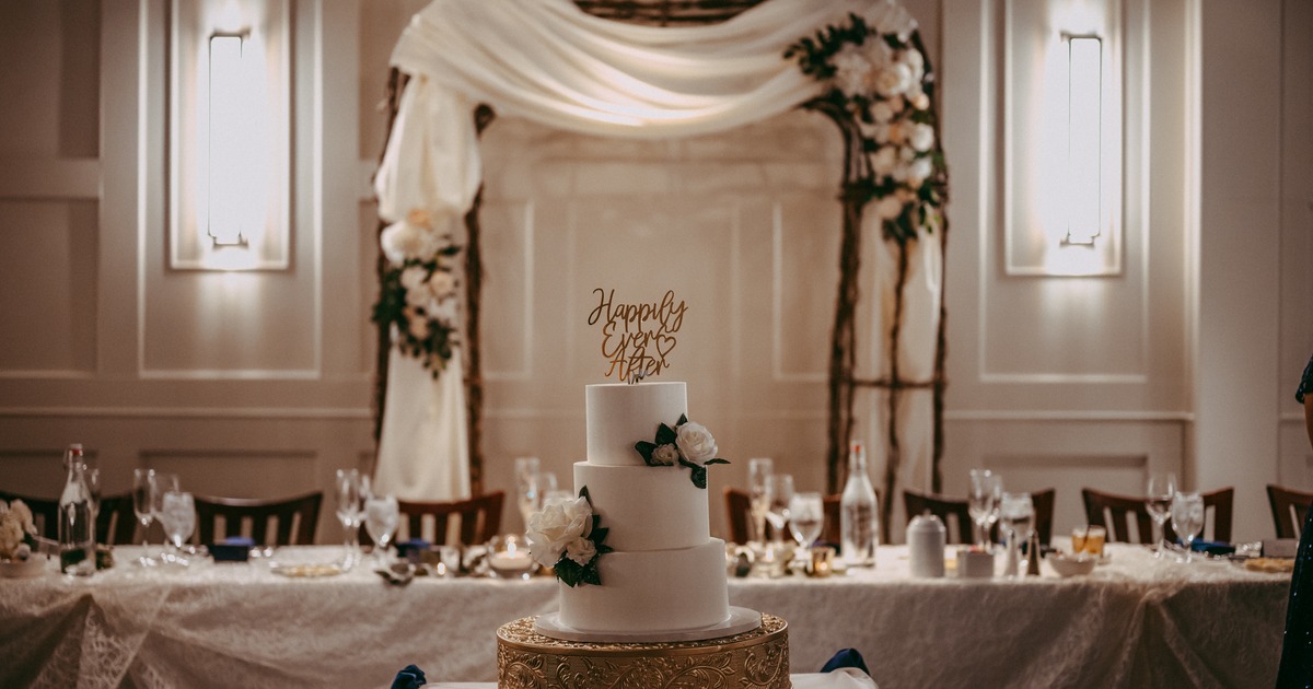 A white wedding cake displayed on a stand in front of a wedding table