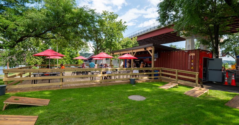 An outdoor shipping-container bar featuring patio seating and lawn games by the river