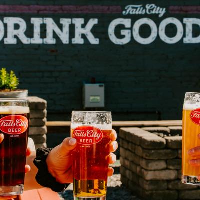 3 people holding glasses of Falls City Beer; a wall in the background displaying the text 