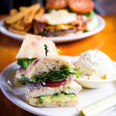 Chicken sandwich on a cibatta roll with lettuce, tomato and avocado inside. Plated next to a serving of potato salad