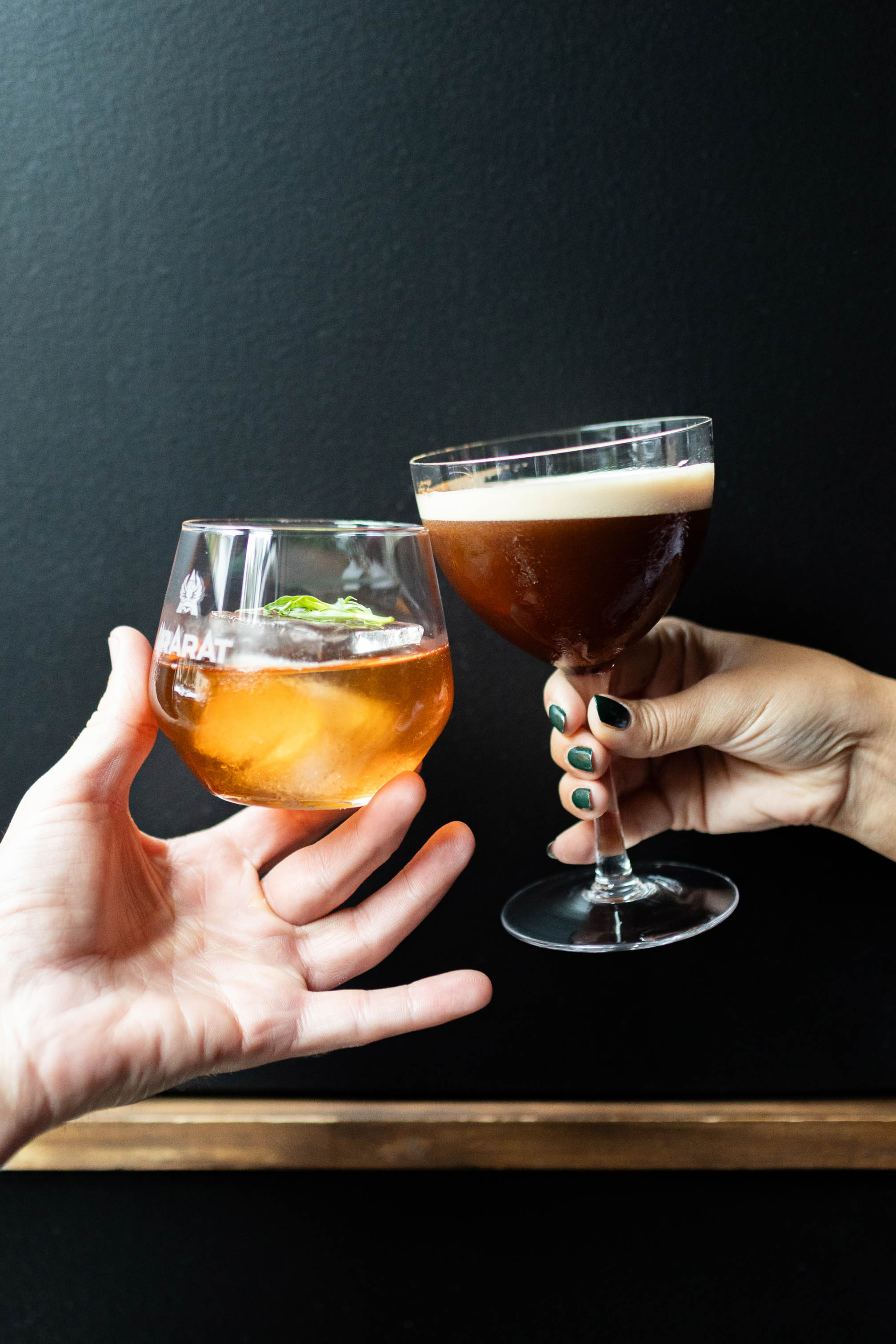 two hands clink cocktails, one an espresso martini and the other an old-fashioned.