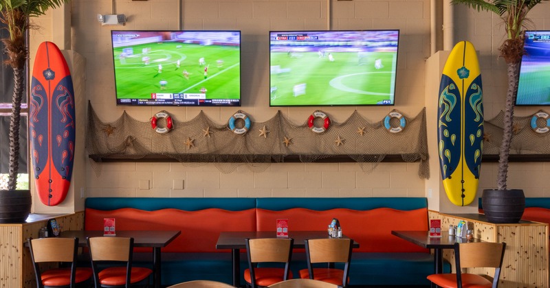 A nautical-themed dining area with surfboards, TVs showing soccer and orange seating.