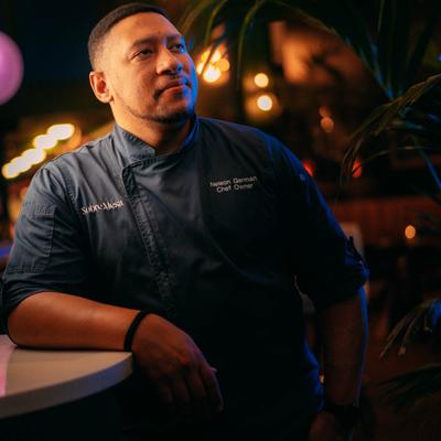 Chef in a blue uniform leaning on a counter against a dimly-lit background.