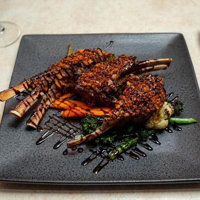 Herb-crusted lamb chops with roasted vegetables and balsamic glaze arranged on dark plate.