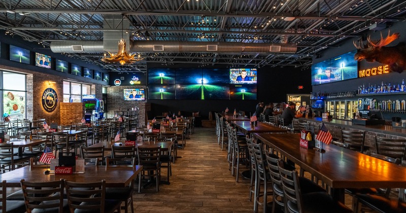 Interior, wooden tables and chairs, a bar with various bottles, large screens showing content