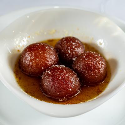 A white bowl holds four Gulab Jamun donuts in a sweet saffron syrup.
