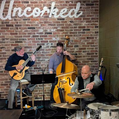 A guitarist, a bassist, and a drummer playing live against a brick wall background.