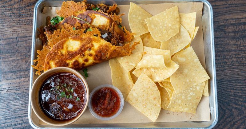 Two birria tacos served with consomme, tortilla chips, and red salsa