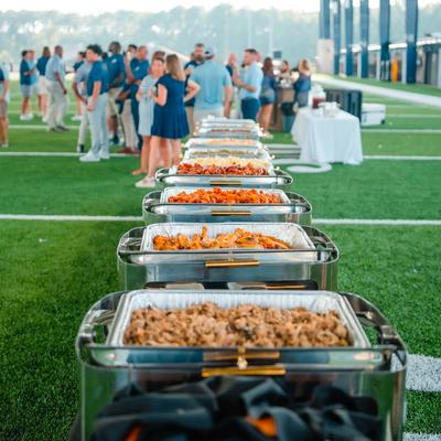 A row of chafing dishes with food lined up on a football field for attendees at an event.