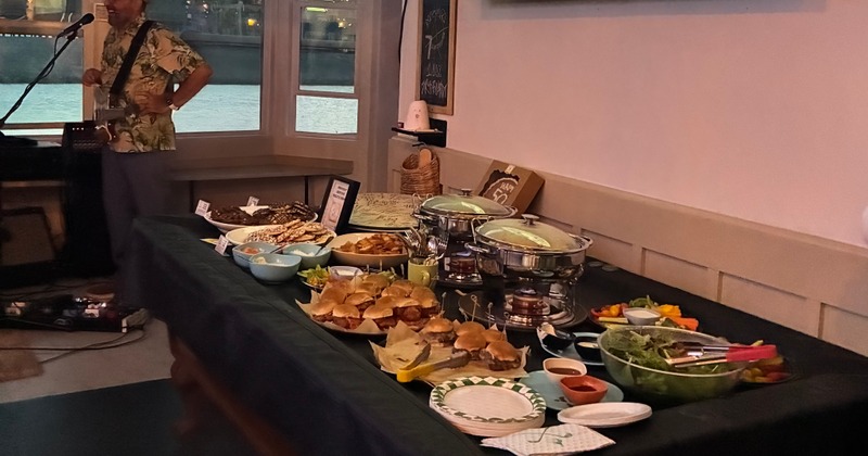 A black table with sliders, vegetable platters, green salad, 2 warming dishes, pita chips, 3 bowls of dips, and cookies
