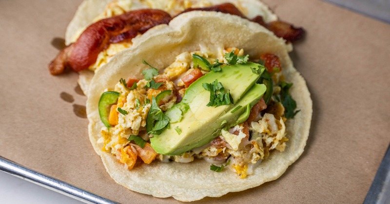 Two breakfast tacos topped with avocado, scrambled eggs, and bacon