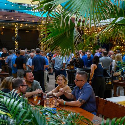 A vibrant outdoor gathering with people at wooden tables under string lights and palm fonds.