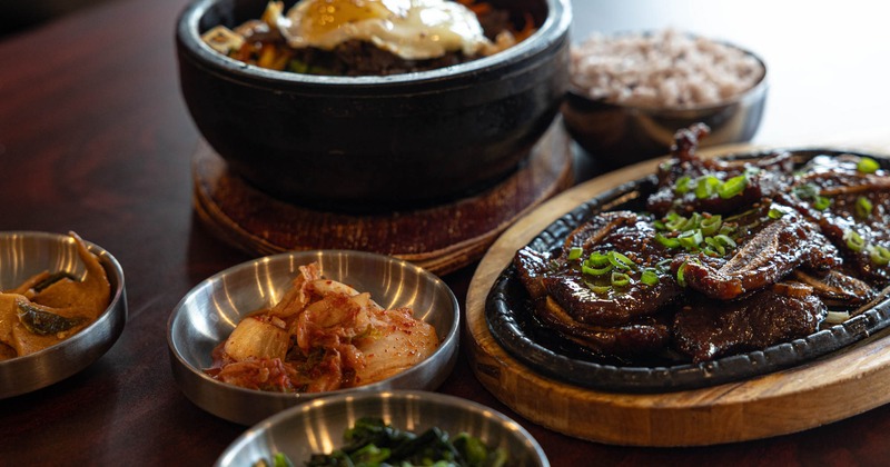 A skillet of Korean short ribs, a stone pot rice bowl, and various side dishes on a table