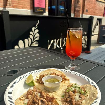 Three tacos on a paper plate with a side of sauce and a lime wedge, accompanied with a drink.