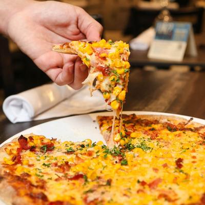A person lifts a  pizza slice topped with corn, cheese, and herbs from a pizza on a plate.