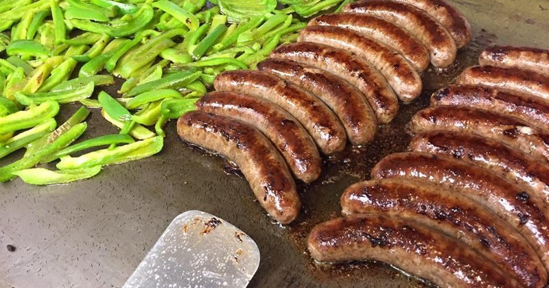 Sausages cooking on a flat-top grill alongside sliced green bell peppers