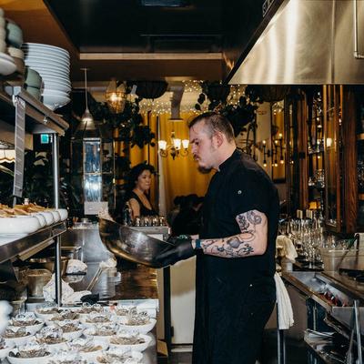 A chef with tattoos mixing ingredients in a metal bowl.