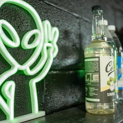 Green alien neon sign on a shelf with a bottle of simple syrup nearby.